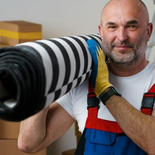 Man mover in uniform carrying big rolled carpet for relocation